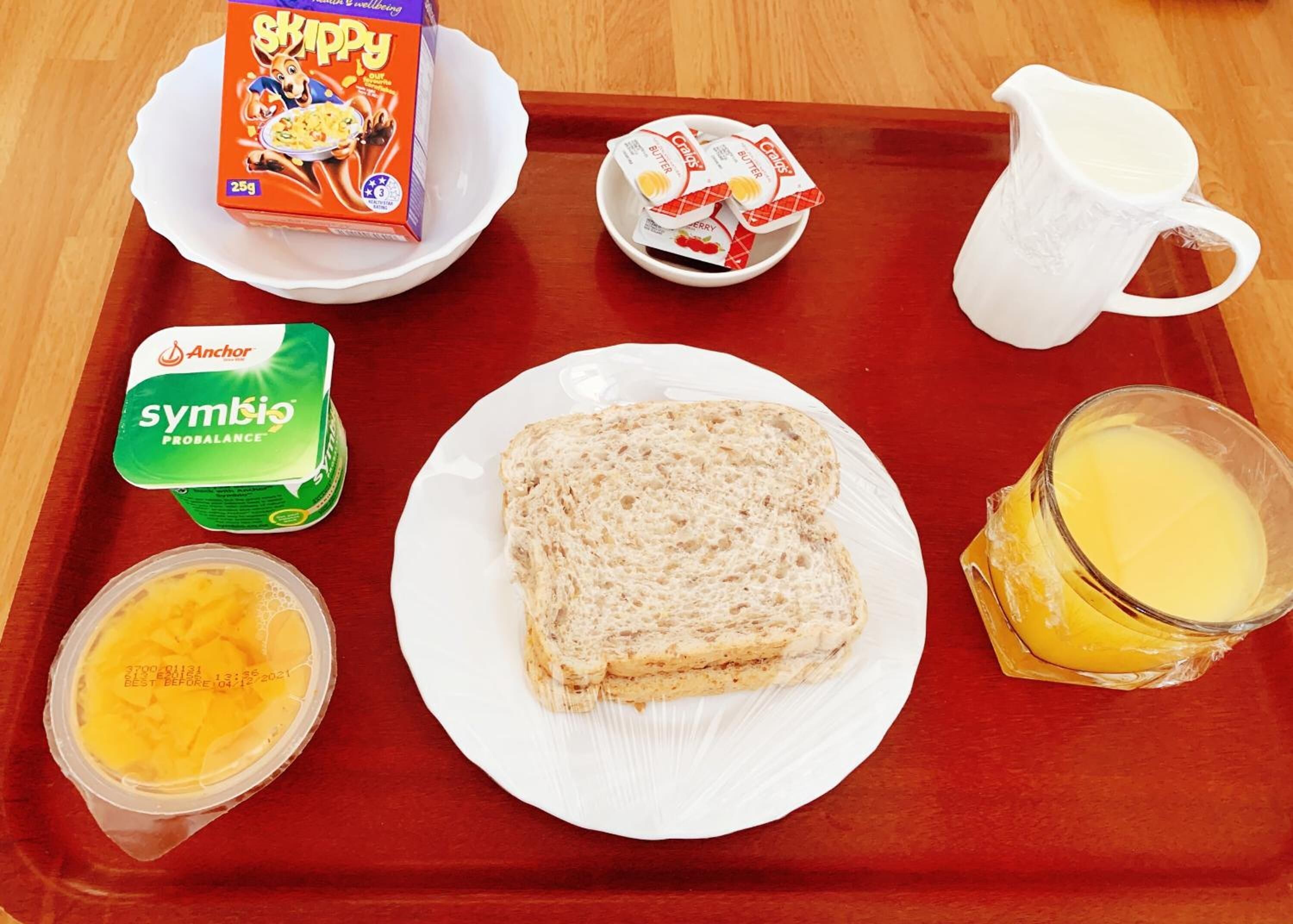 Uma bandeja com um pote de manteiga de amendoim Skippy, um recipiente de iogurte Symbio ProBalance, um copo de suco de laranja, uma fatia de pão e uma jarra de leite.