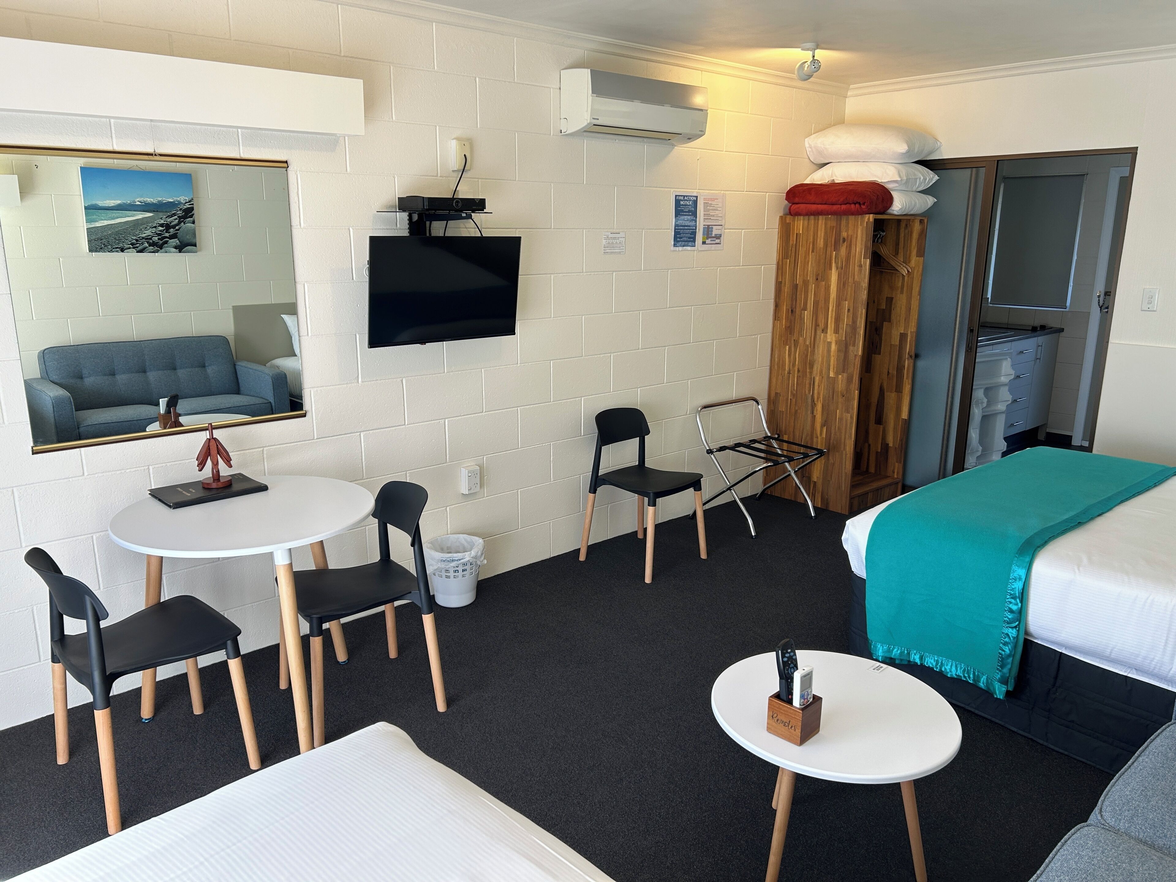 A hotel room with a bed, a desk with a chair, a small table with a chair, a TV mounted on the wall, and a window with a view of a beach.