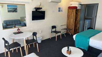 A hotel room with a bed, a desk with a chair, a small table with a chair, a TV mounted on the wall, and a window with a view of a beach.