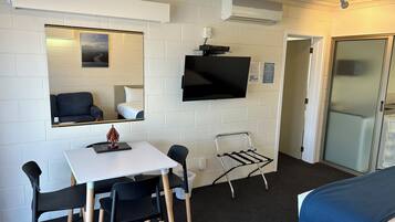 A compact hotel room with a white brick wall, a flat-screen TV mounted above a sofa, a small table with chairs, and a bed with a blue cover.