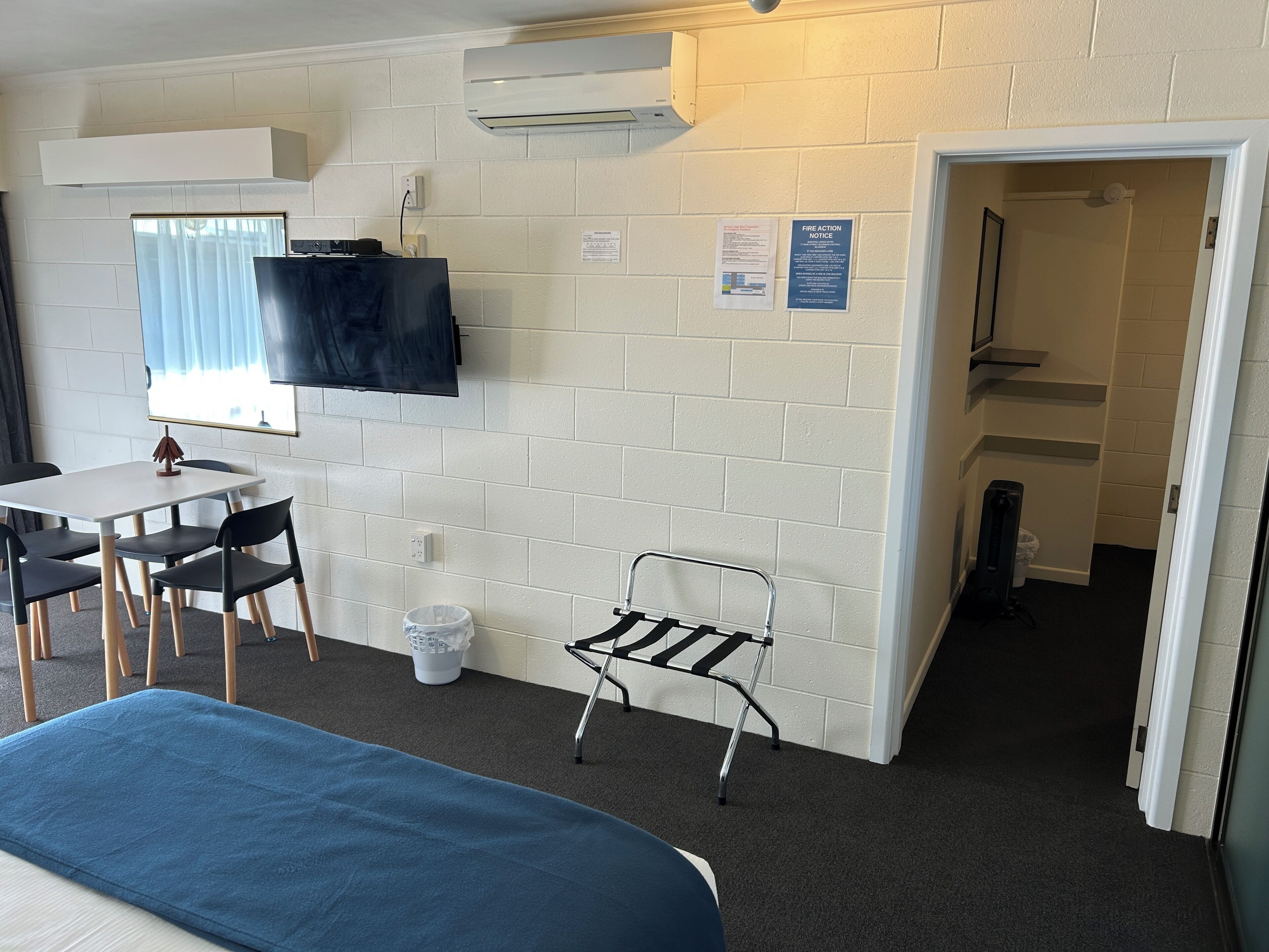 A room with a bed, a desk with chairs, a TV mounted on the wall, and a fire action notice.