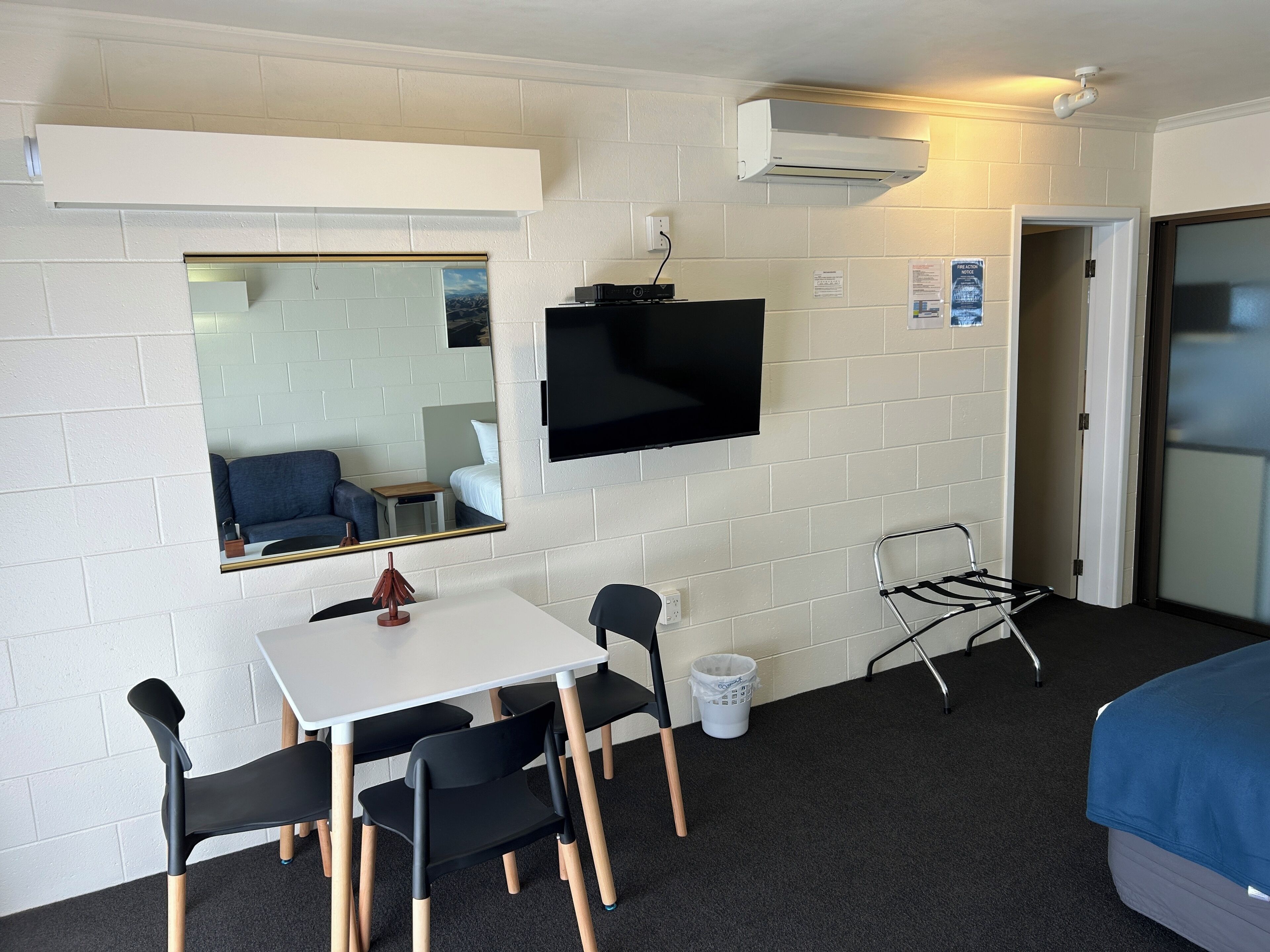 A compact hotel room with a single bed, a small table with chairs, a TV mounted on the wall, and a mirror reflecting the room.