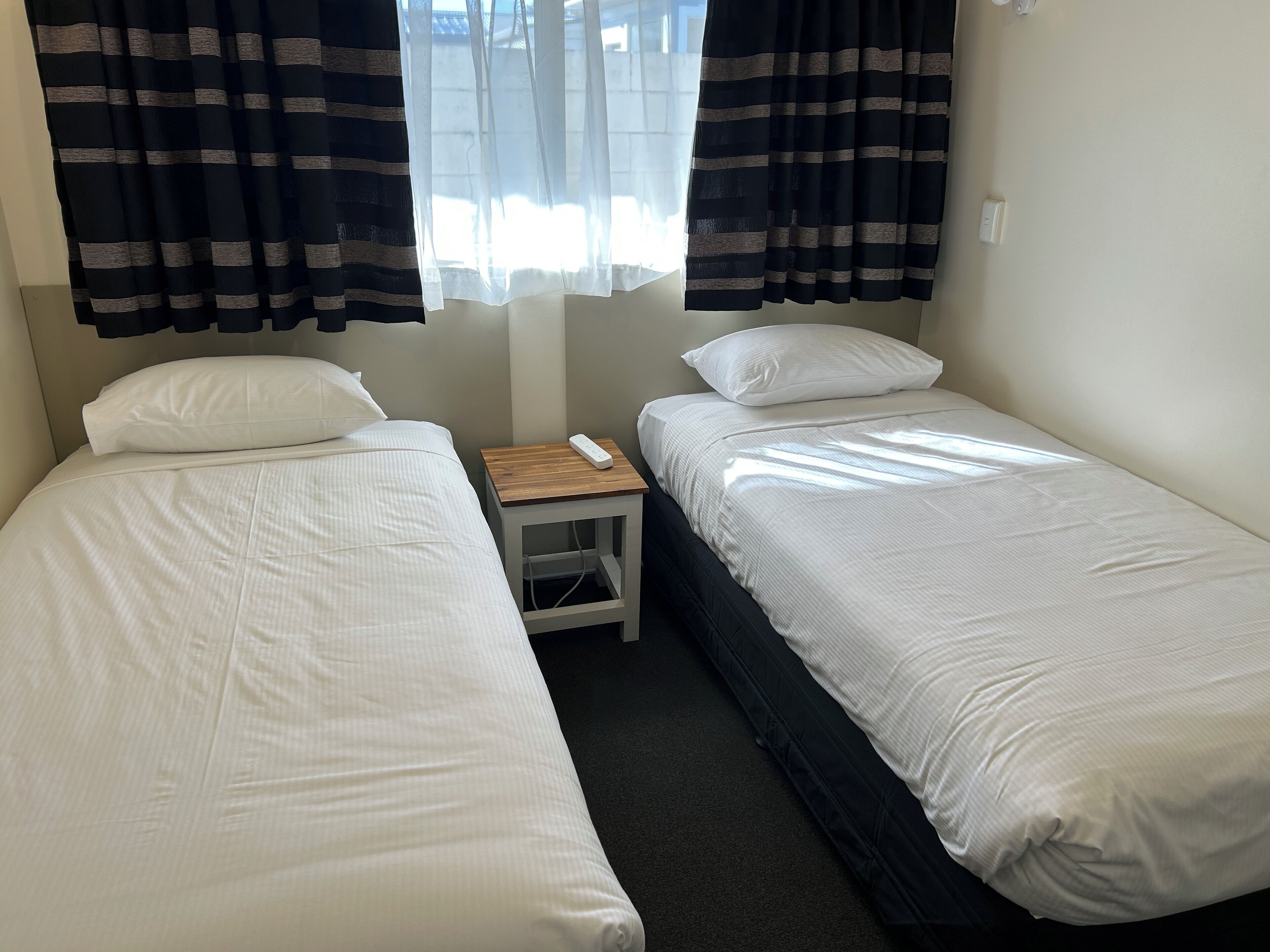 Een kamer met twee eenpersoonsbedden, een klein houten nachtkastje en een raam met gordijnen.