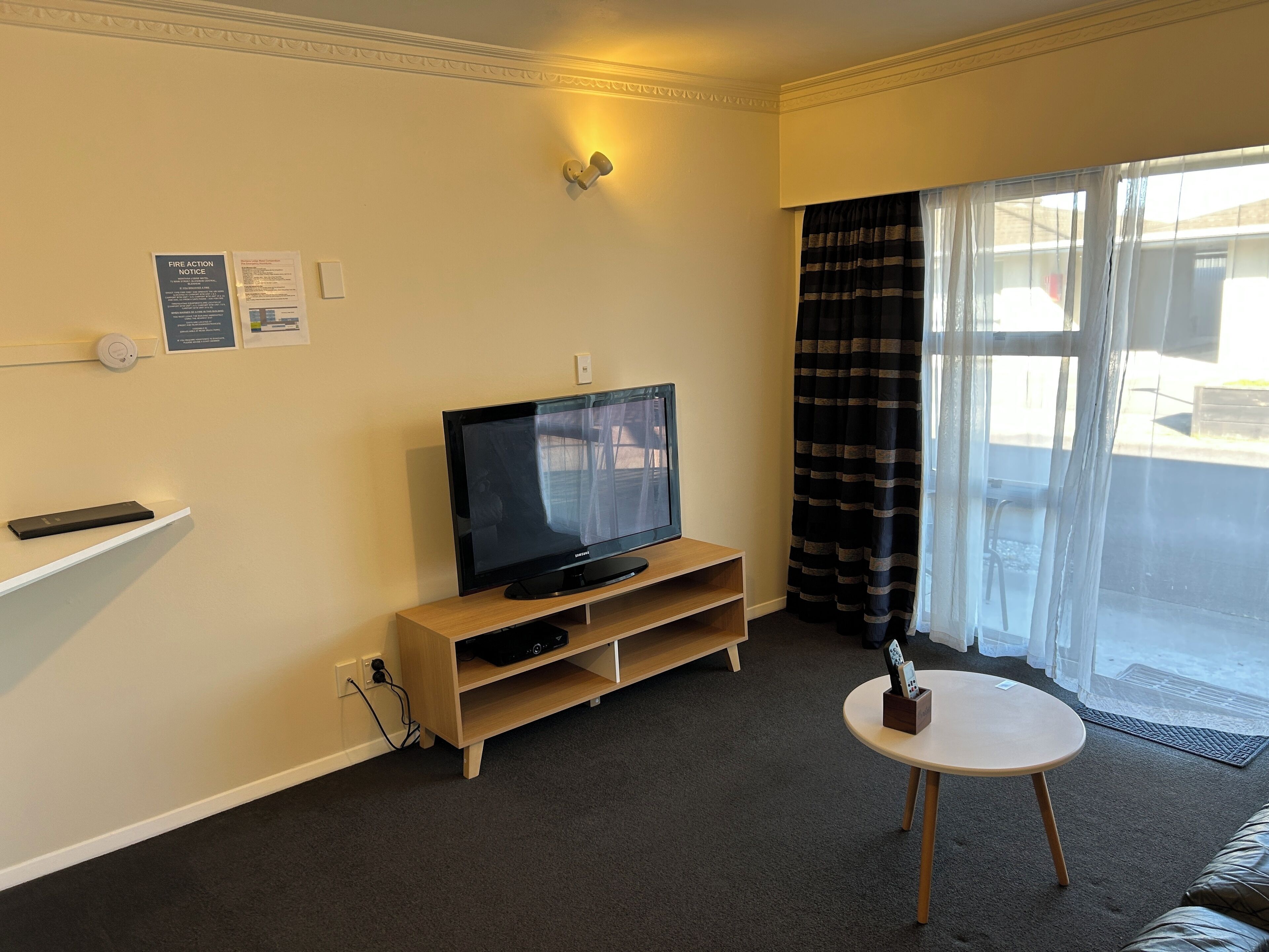 A living room with a flat-screen TV on a wooden stand, a small round table, and a large window with curtains.