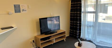 A living room with a flat-screen TV on a wooden stand, a small round table, and a large window with curtains.