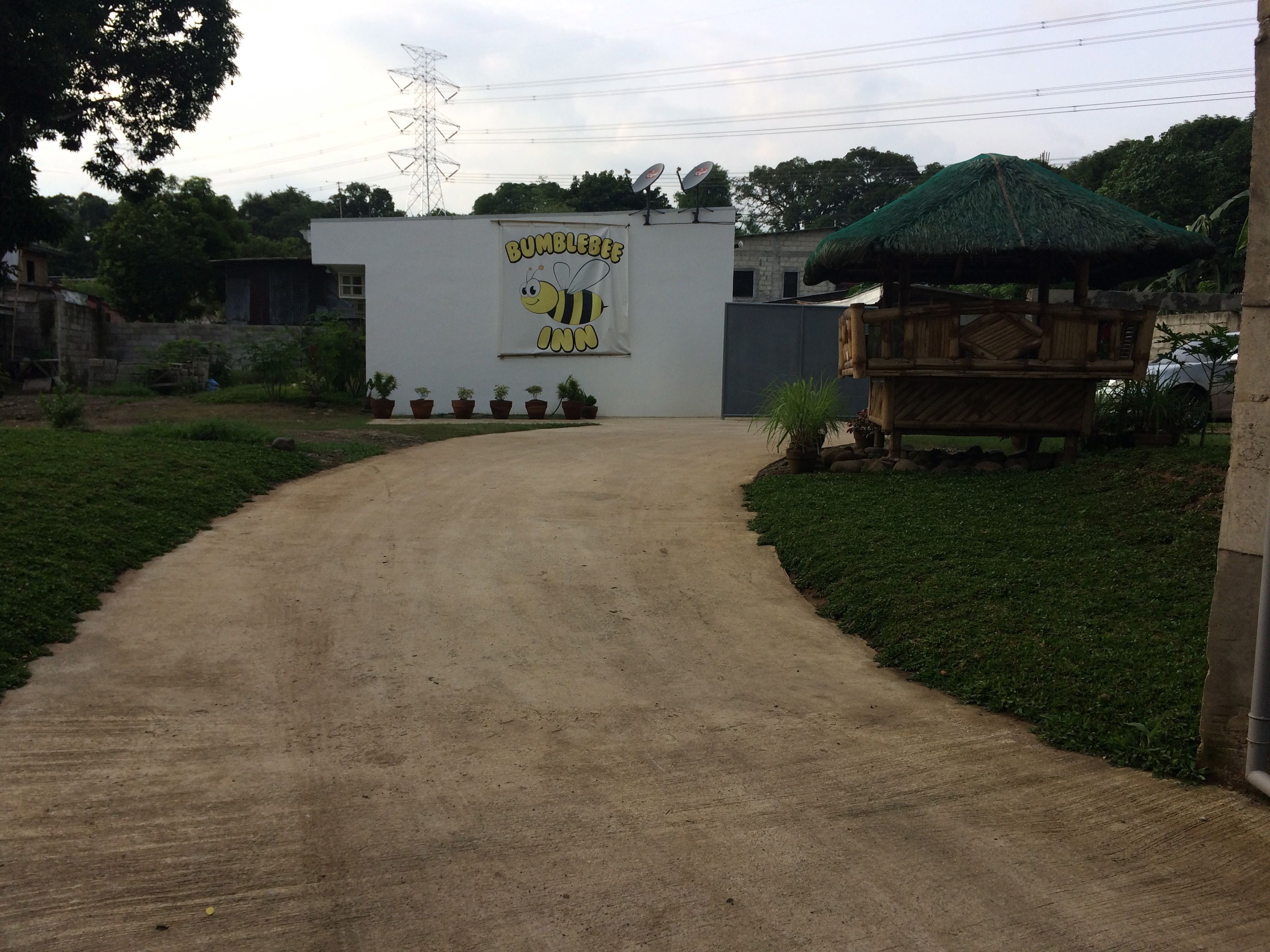 A dirt road leading to a building with a 'Bumblebee Inn' sign, surrounded by greenery and a thatched-roof structure.