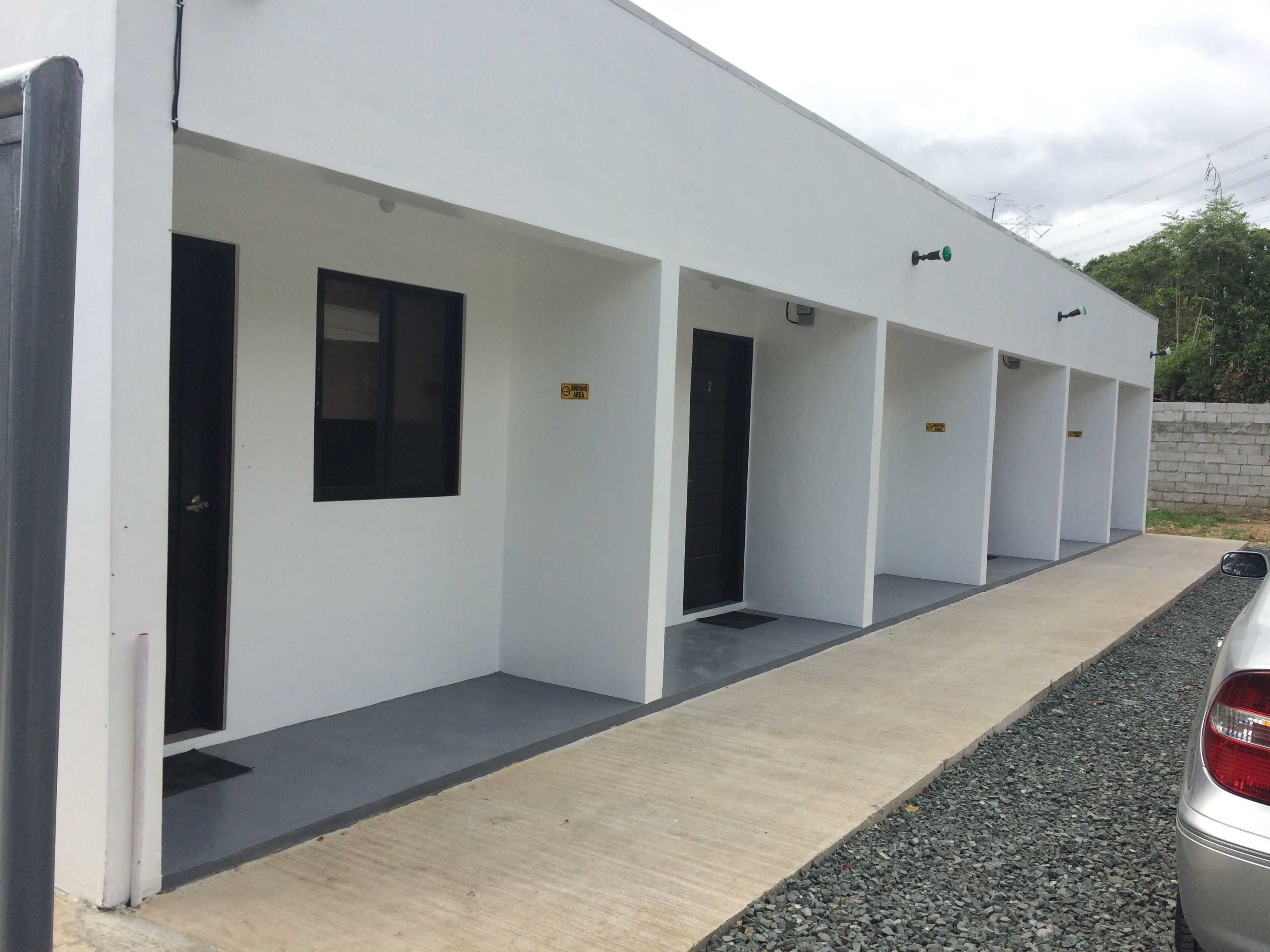 A row of single-room hotel units with individual doorways, a concrete pathway, and a parked car.