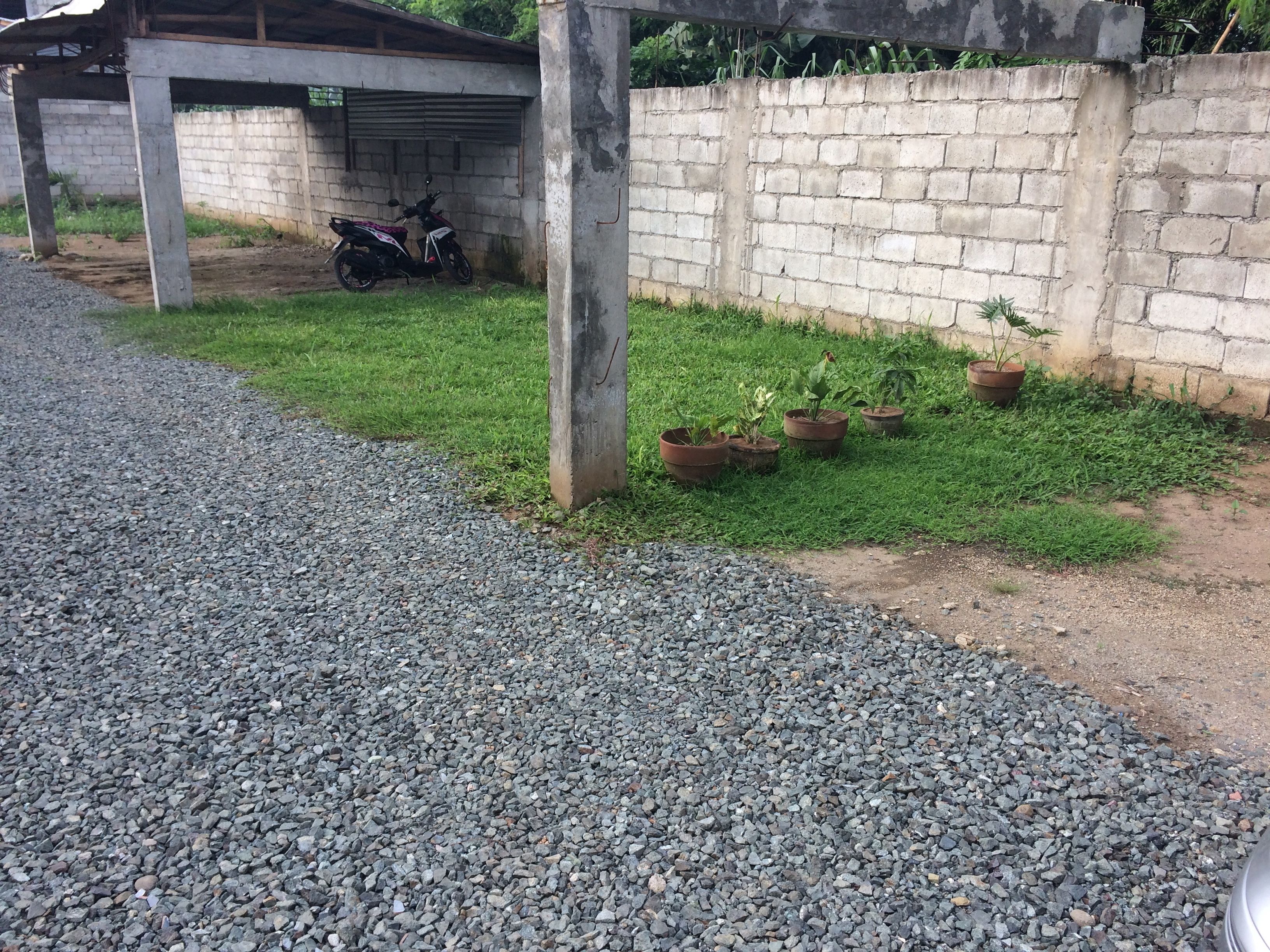 Un camino de grava que conduce a un área de estacionamiento techada, con un muro de concreto y plantas en macetas.