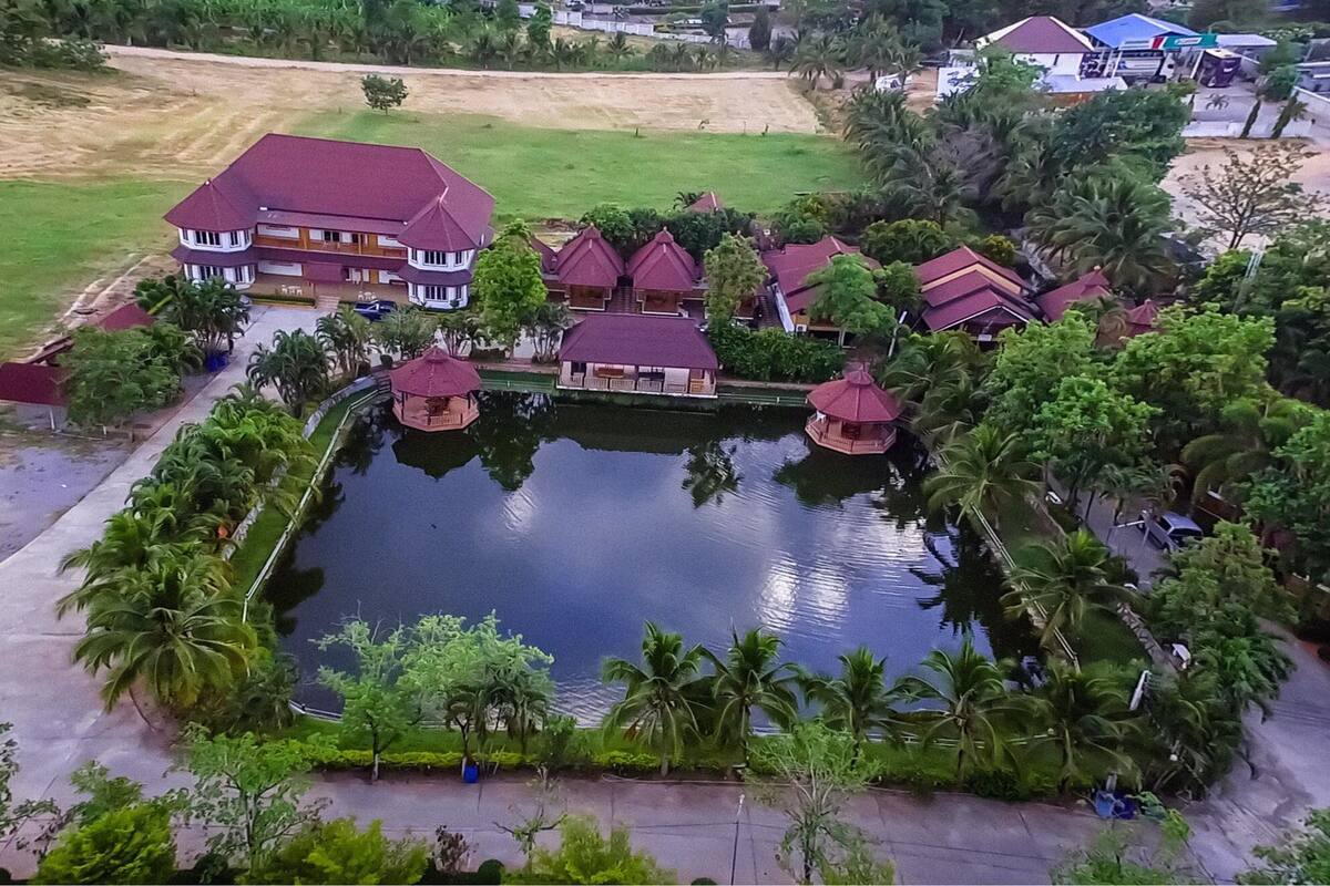 An aerial view of a resort with a central pond, multiple buildings with red roofs, and surrounding palm trees.