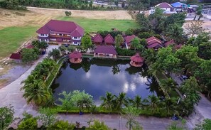 An aerial view of a resort with a central pond, multiple buildings with red roofs, and surrounding palm trees.
