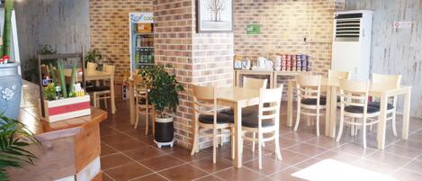 Una cafetería con paredes de ladrillo, mesas y sillas de madera. Hay plantas en macetas y un área de autoservicio para bebidas.