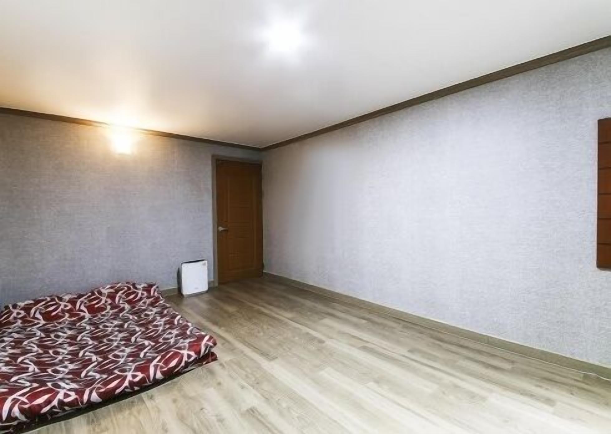 A bedroom with a bed, a wooden floor, a door, and a wall-mounted light fixture.