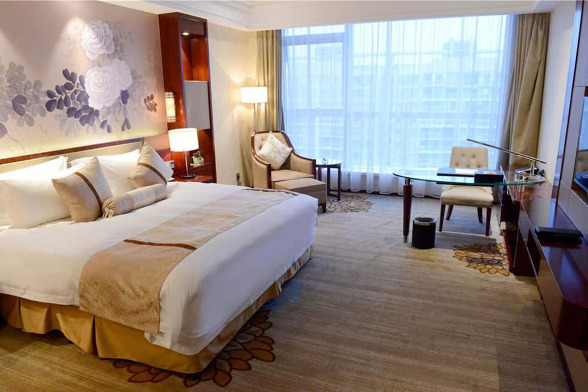 A hotel room with a large bed, a desk, a chair, and a view of the city through large windows.