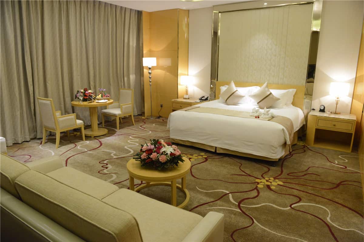 A hotel room with a large bed, a sofa, a small table with flowers, a desk, and a lamp.