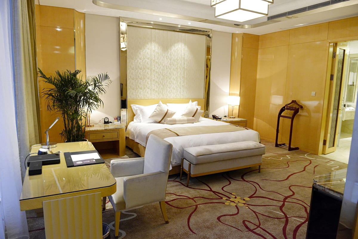 A hotel room with a bed, bedside tables, a desk, a chair, a lamp, and a potted plant.