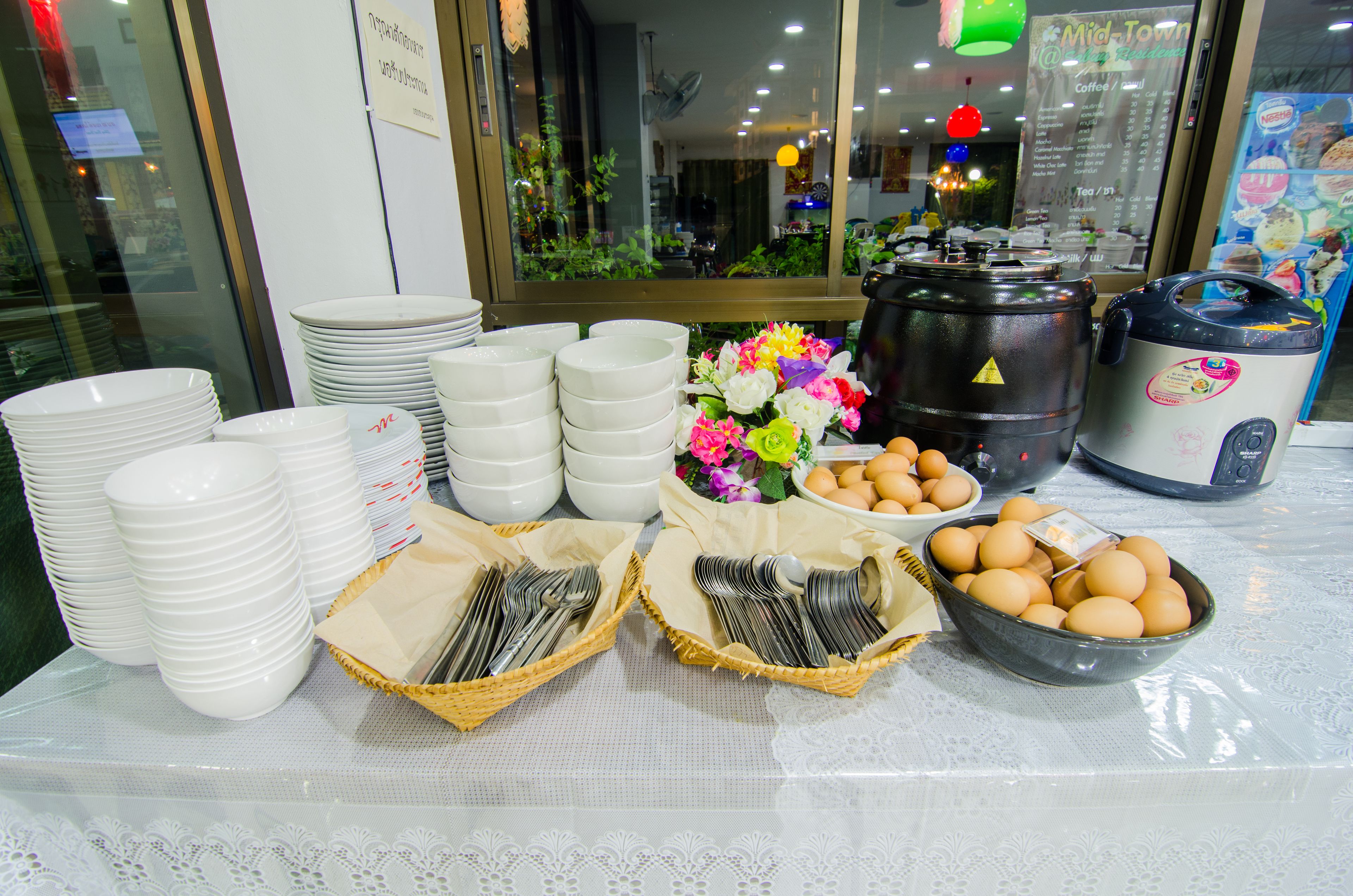 A table with stacks of white plates, bowls, and baskets of utensils, along with a coffee maker and a bowl of eggs.