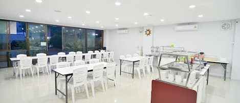 Una habitación amplia con mesas y sillas blancas, una zona de buffet y ventanas grandes.