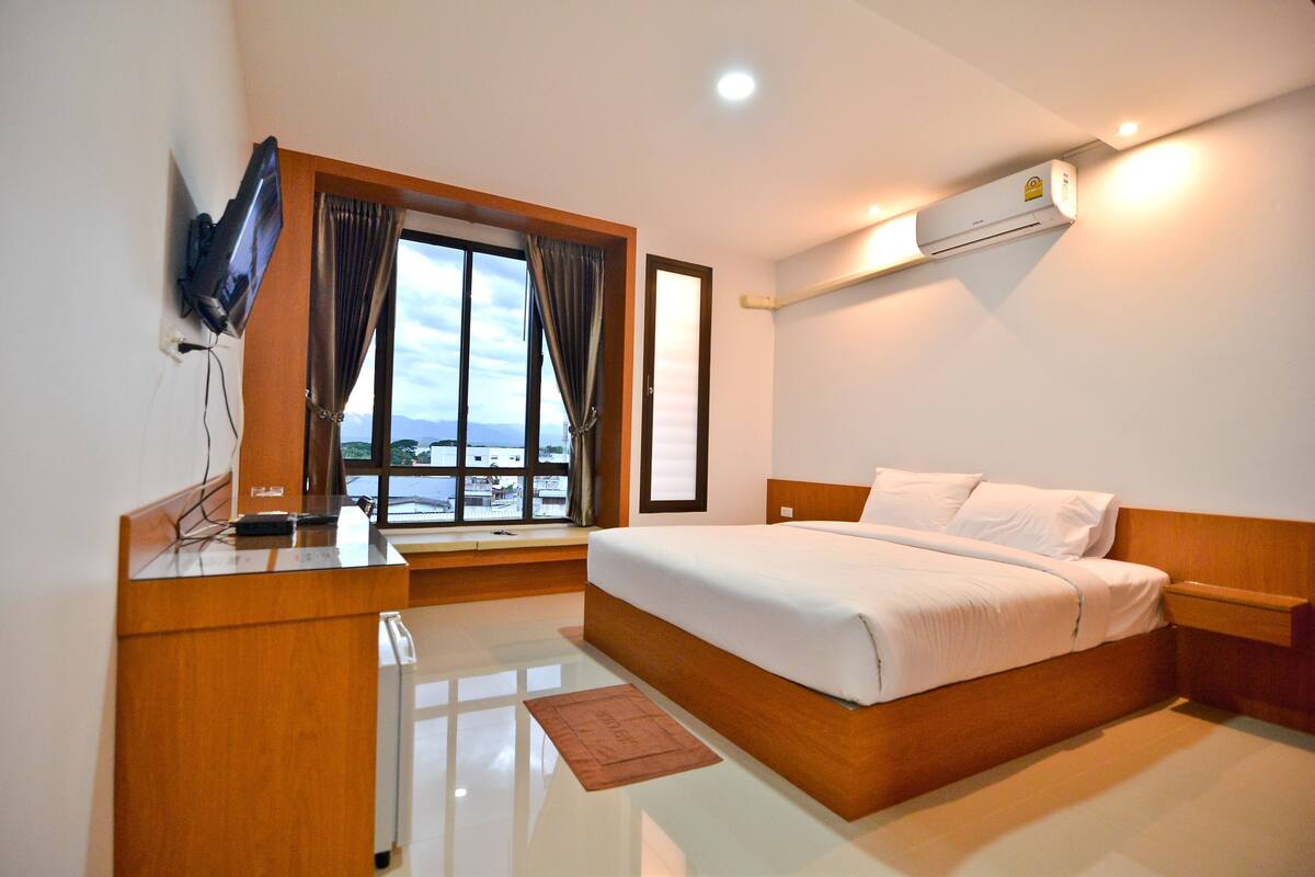 Una habitación de hotel con cama, escritorio, televisión, ventana con cortinas y aire acondicionado.