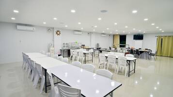 Una amplia sala de conferencias bien iluminada, con mesas y sillas blancas, unidades de aire acondicionado y un reloj en la pared.