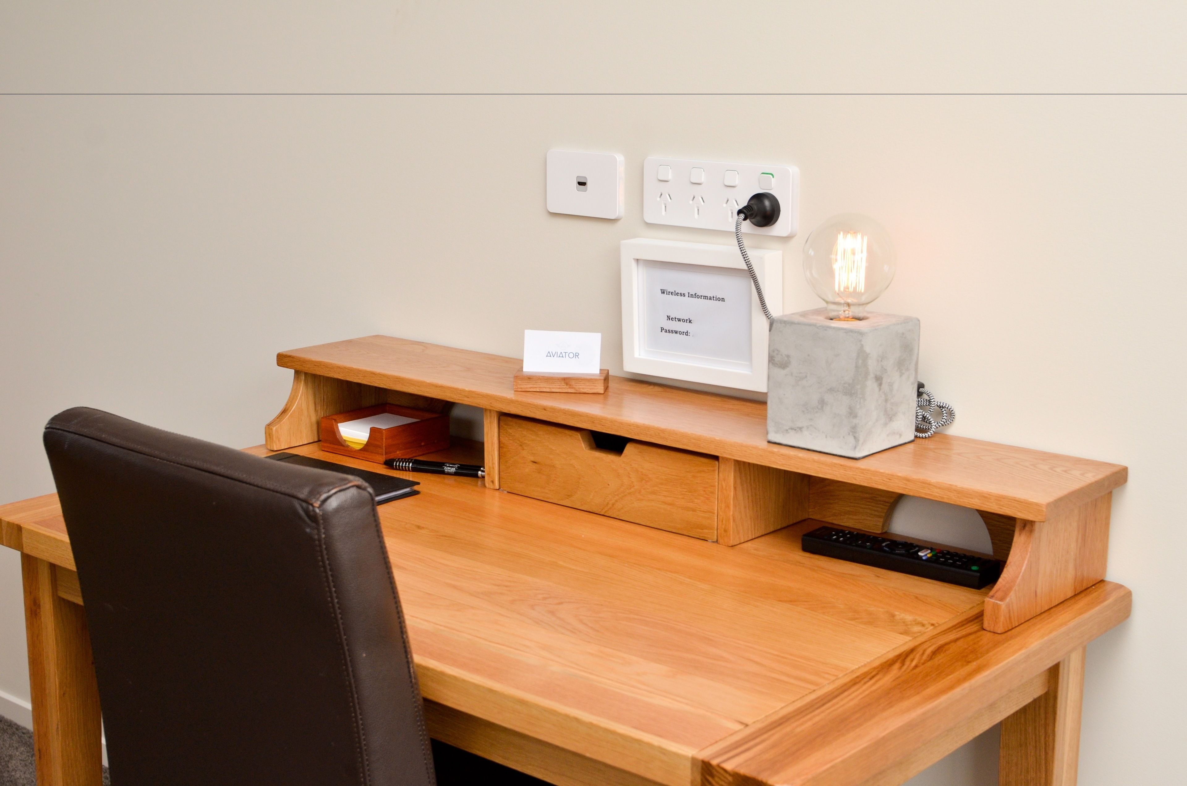 Ein Holztisch mit einem Laptop, einem Schild mit der Aufschrift 'AVIATOR', einem gerahmten Bild, einer Schreibtischlampe und einer Steckdose.