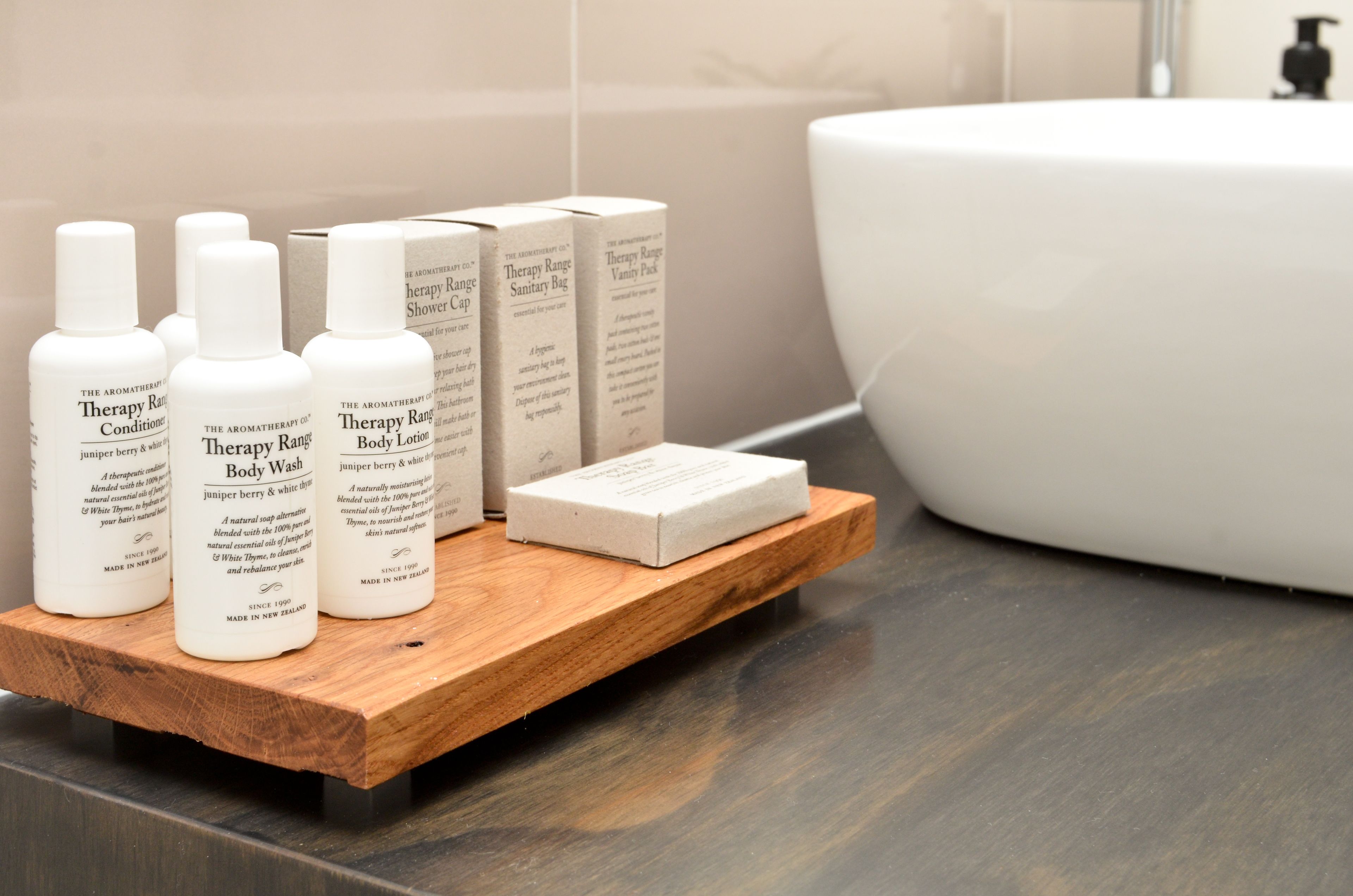 Estante de madera con botellas de jabón corporal 'Therapy Range' y una caja de bolsas sanitarias 'Therapy Range' en un baño.