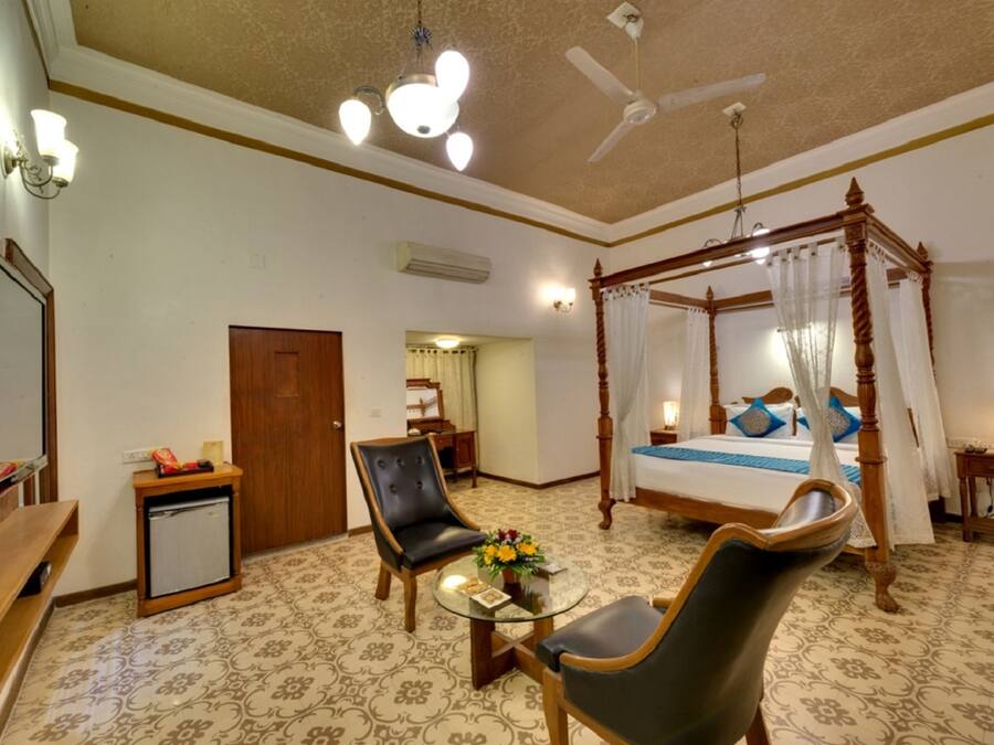 A hotel room with a four-poster bed, a sitting area with a glass table, a ceiling fan, and a television.