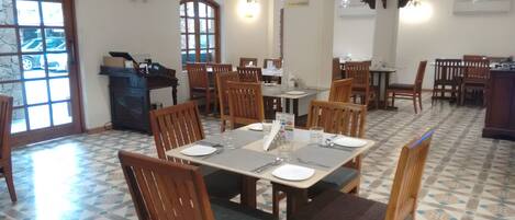 A dining area with wooden tables and chairs, a tiled floor, and a view of the outside through a window.