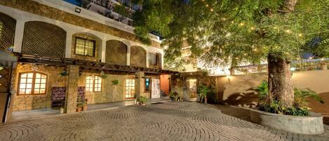 A courtyard with cobblestone paving, a stone building with arched windows, and a tree with string lights.