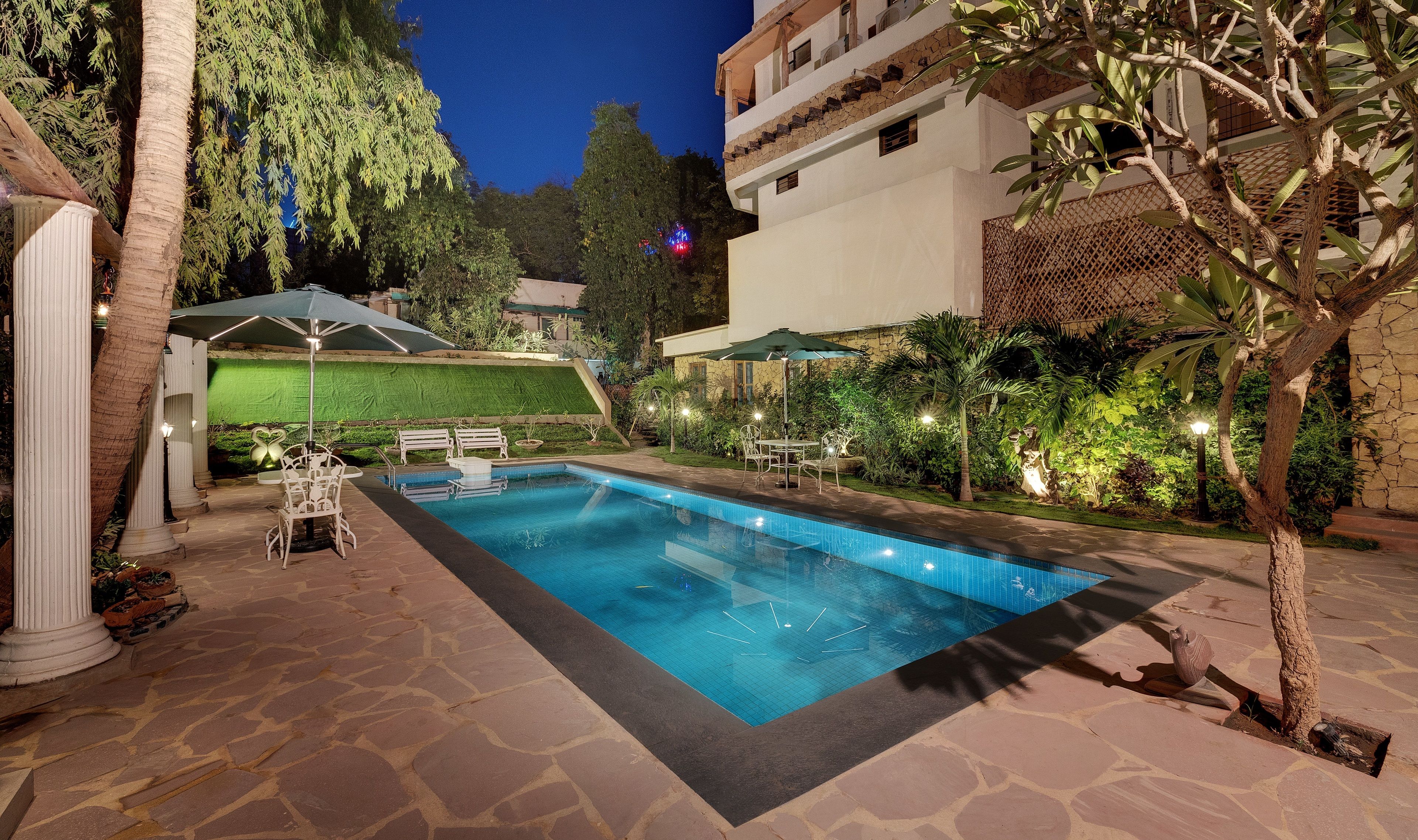 A swimming pool with stone tiles, surrounded by a patio with chairs and umbrellas, and a building with balconies in the background.