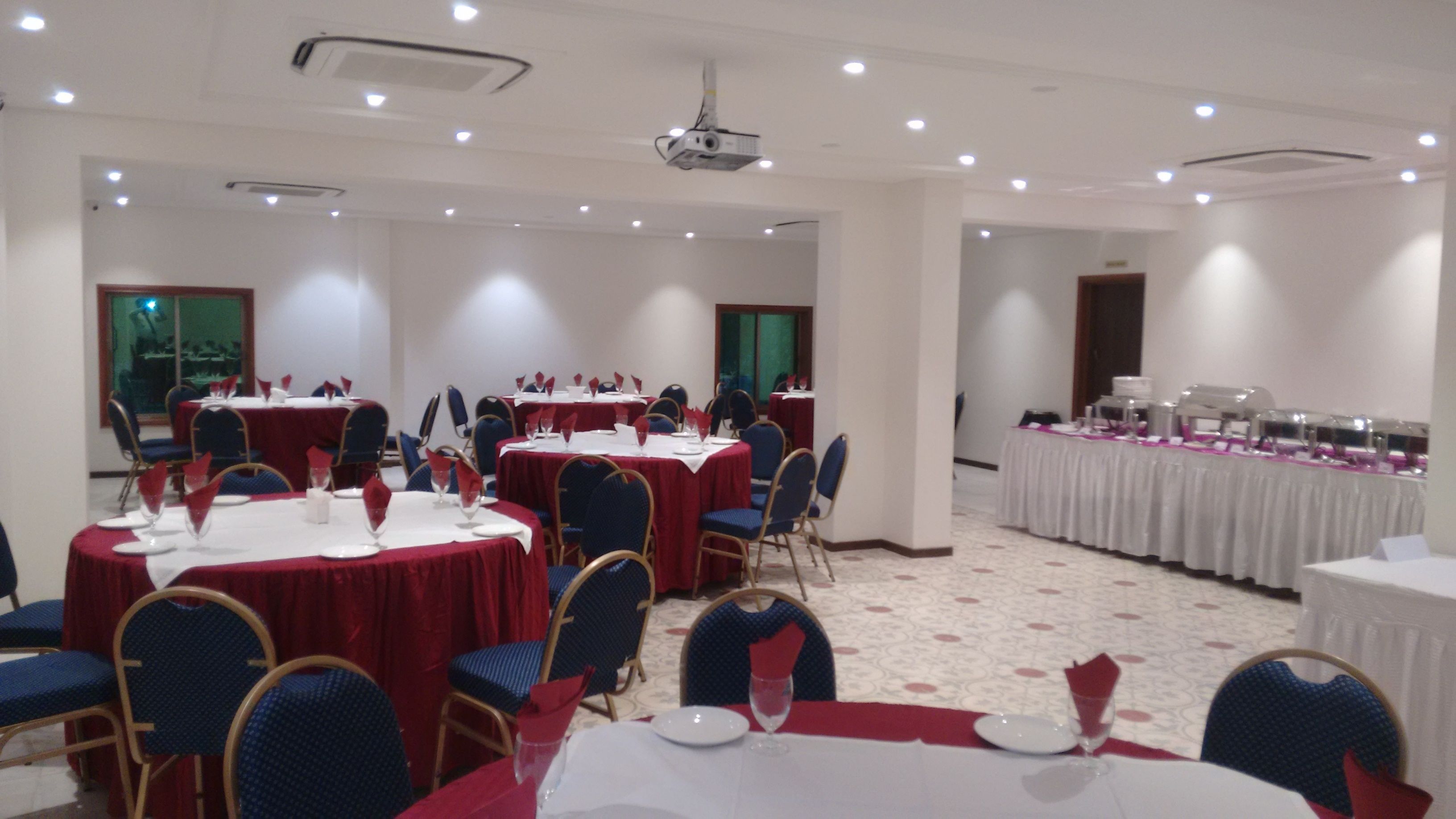 A banquet hall with round tables covered in white tablecloths and red napkins, blue chairs, and a projector on the ceiling.
