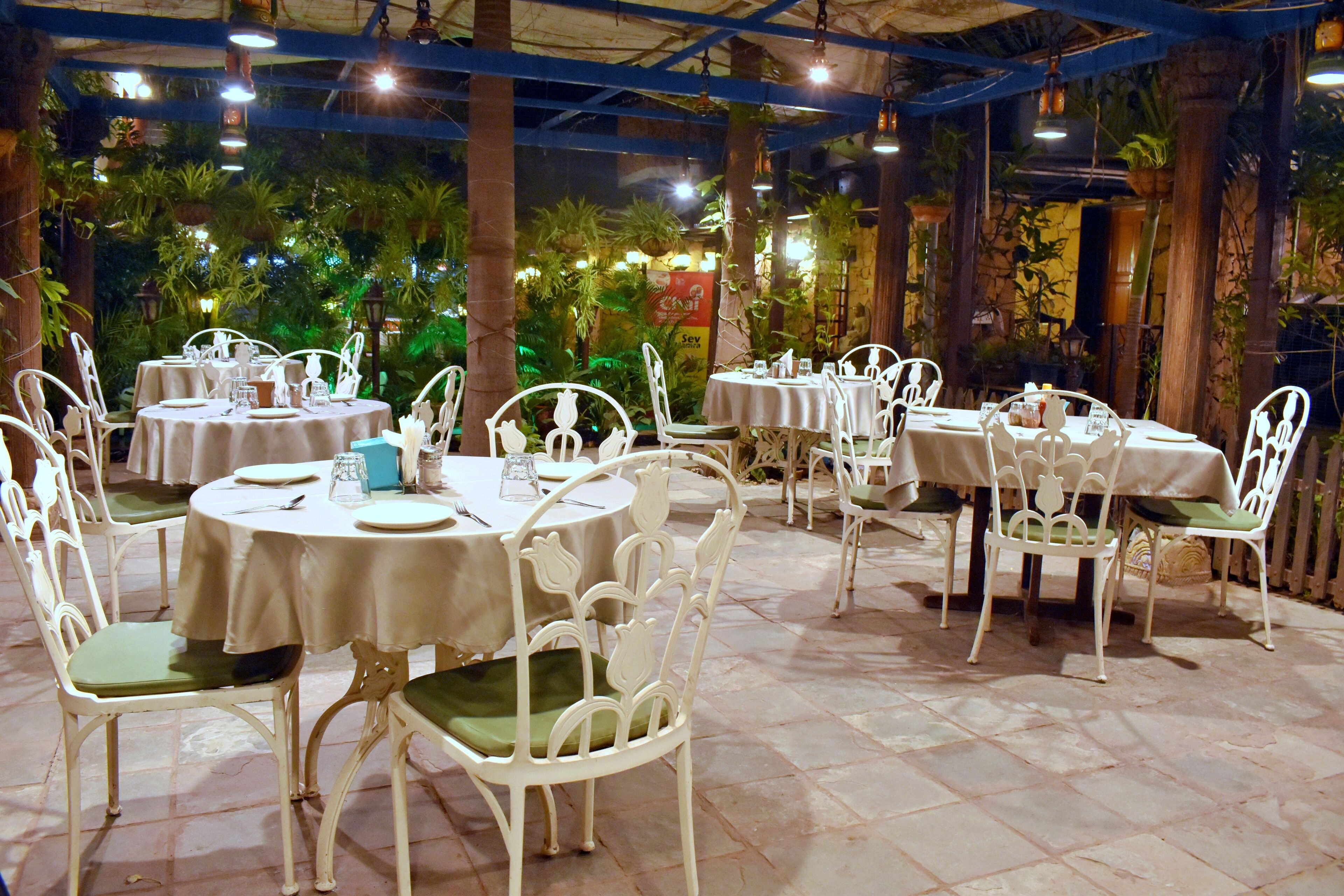 Un restaurante al aire libre con mesas redondas y sillas blancas, rodeado de vegetación.