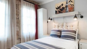 Un dormitorio con cama, mesita de noche, lámpara de pared, un cuadro y ventana con cortinas.