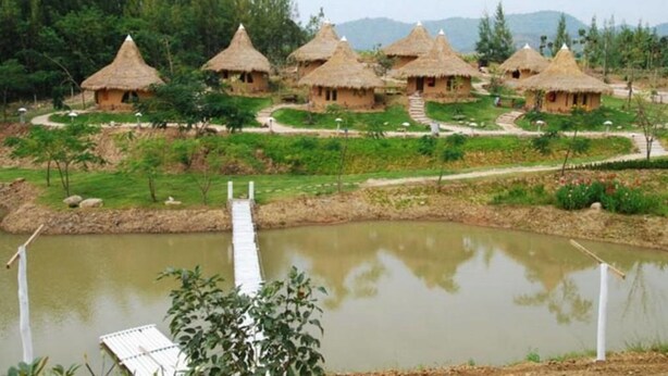 A series of thatched-roof huts arranged in a semi-circle with a wooden bridge over a pond. - Ban Rai Pornsawan (Suan Phueng)