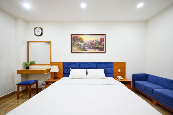 Une chambre d’hôtel bien rangée, avec un lit, un canapé bleu, un bureau avec une lampe, une petite plante et un tableau accroché au mur.