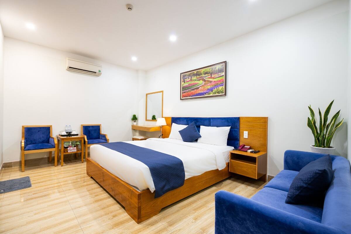 Une chambre d’hôtel avec un lit en bois, des chaises bleues, un canapé, une table de chevet, une lampe, une plante en pot et un tableau au mur.