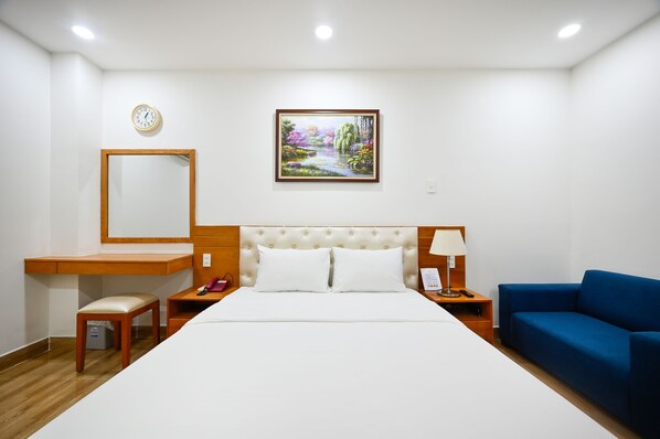 Habitación de hotel con una cama grande, un escritorio, una silla, un sofá azul, una mesita de noche, una lámpara, un reloj y un cuadro en la pared.