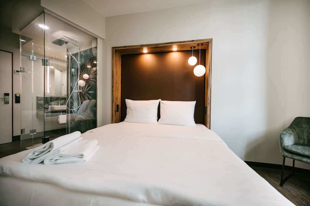A hotel room with a large bed, a glass shower enclosure, a wooden headboard, and a green armchair.
