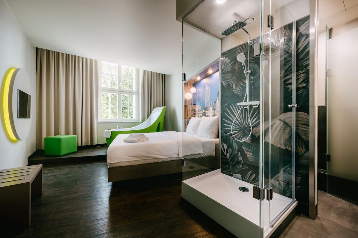 A modern hotel room with a bed, a green chair, a glass shower enclosure, and a large window.