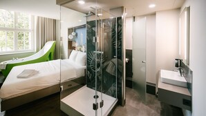 A modern bedroom with a glass-enclosed shower, a bed with a green accent, and a bathroom with a large mirror and sink. - Travel24 Hotel (Leipzig)