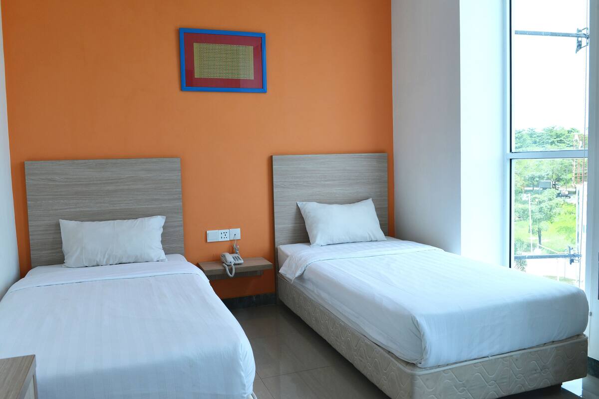 A hotel room with two beds, a small bedside table with a phone, and a window with a view of greenery.