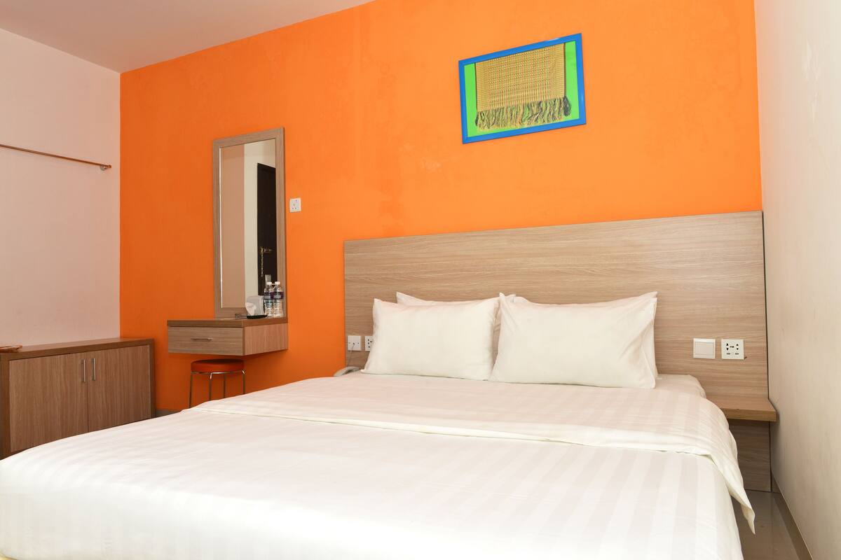 A hotel room with a bed, a wooden headboard, a mirror, a small desk, and an orange wall.