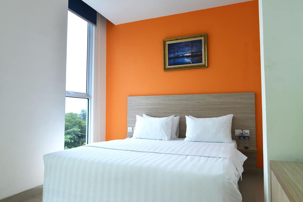 A hotel room with a large bed, a nightstand, a framed picture on the wall, and a window providing a view of greenery.