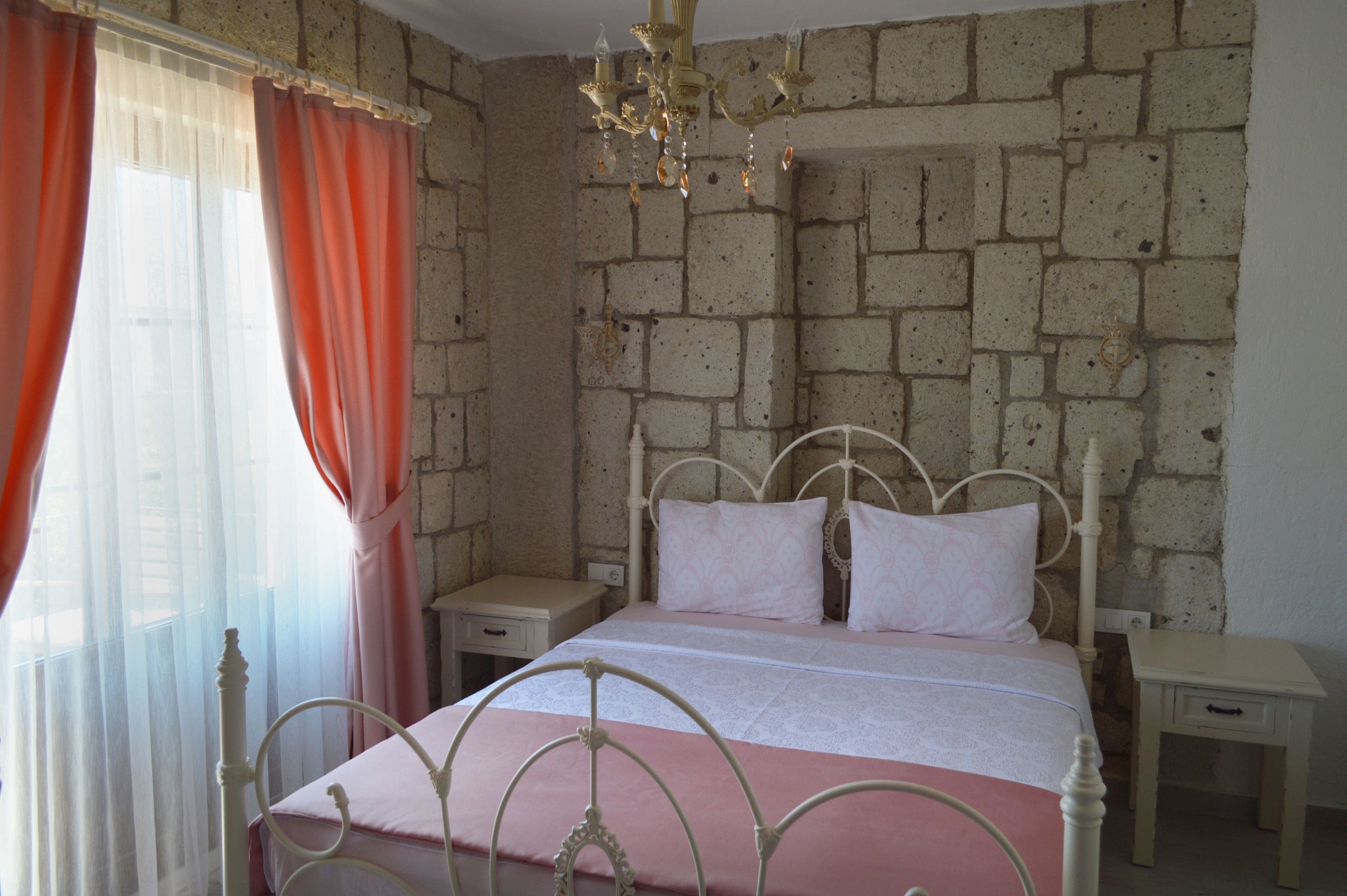 A bedroom with a brick wall, a metal bed frame, a chandelier, and a window with curtains.