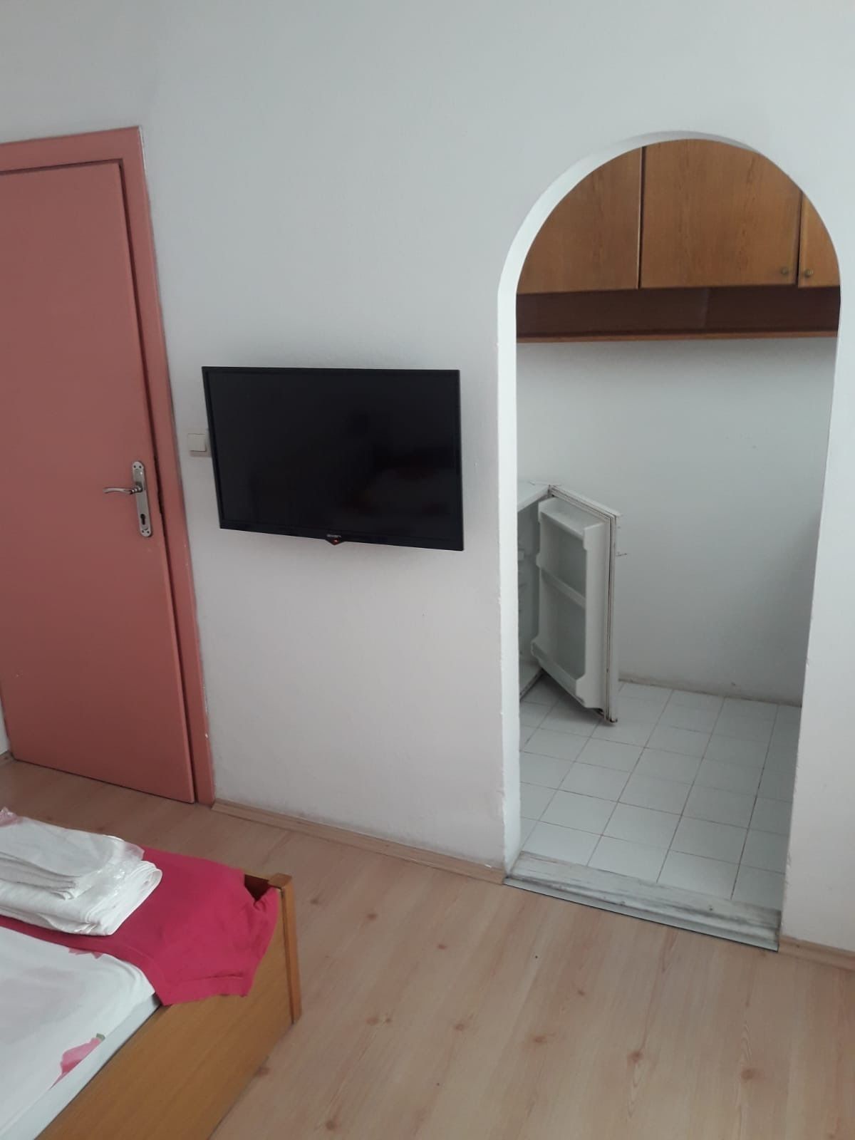 A bedroom with a bed, a TV mounted on the wall, a pink door, and an arched doorway leading to a bathroom with a radiator.