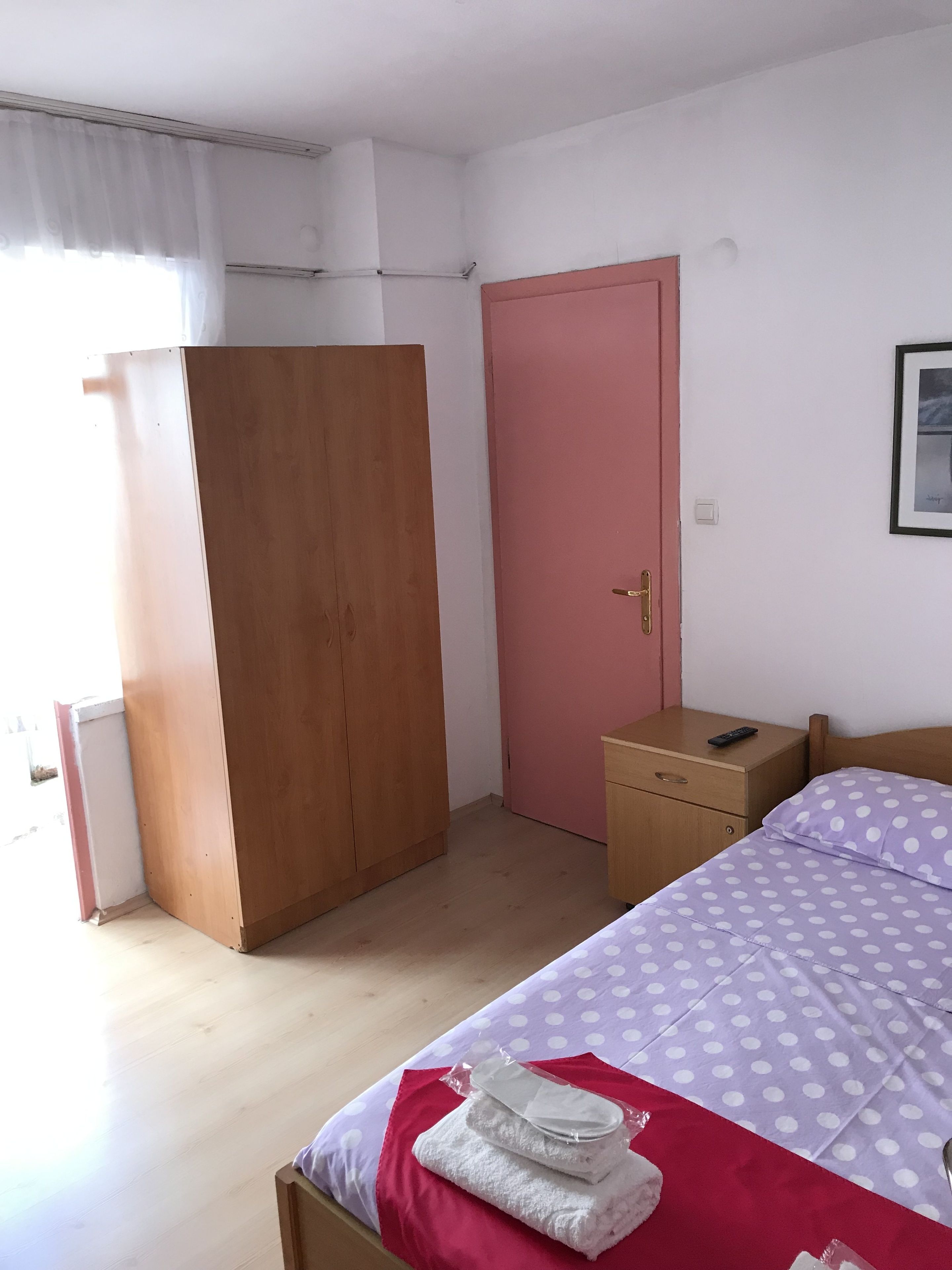 A bedroom with a bed, a wooden wardrobe, a pink door, a bedside table with a remote control, and a framed picture on the wall.