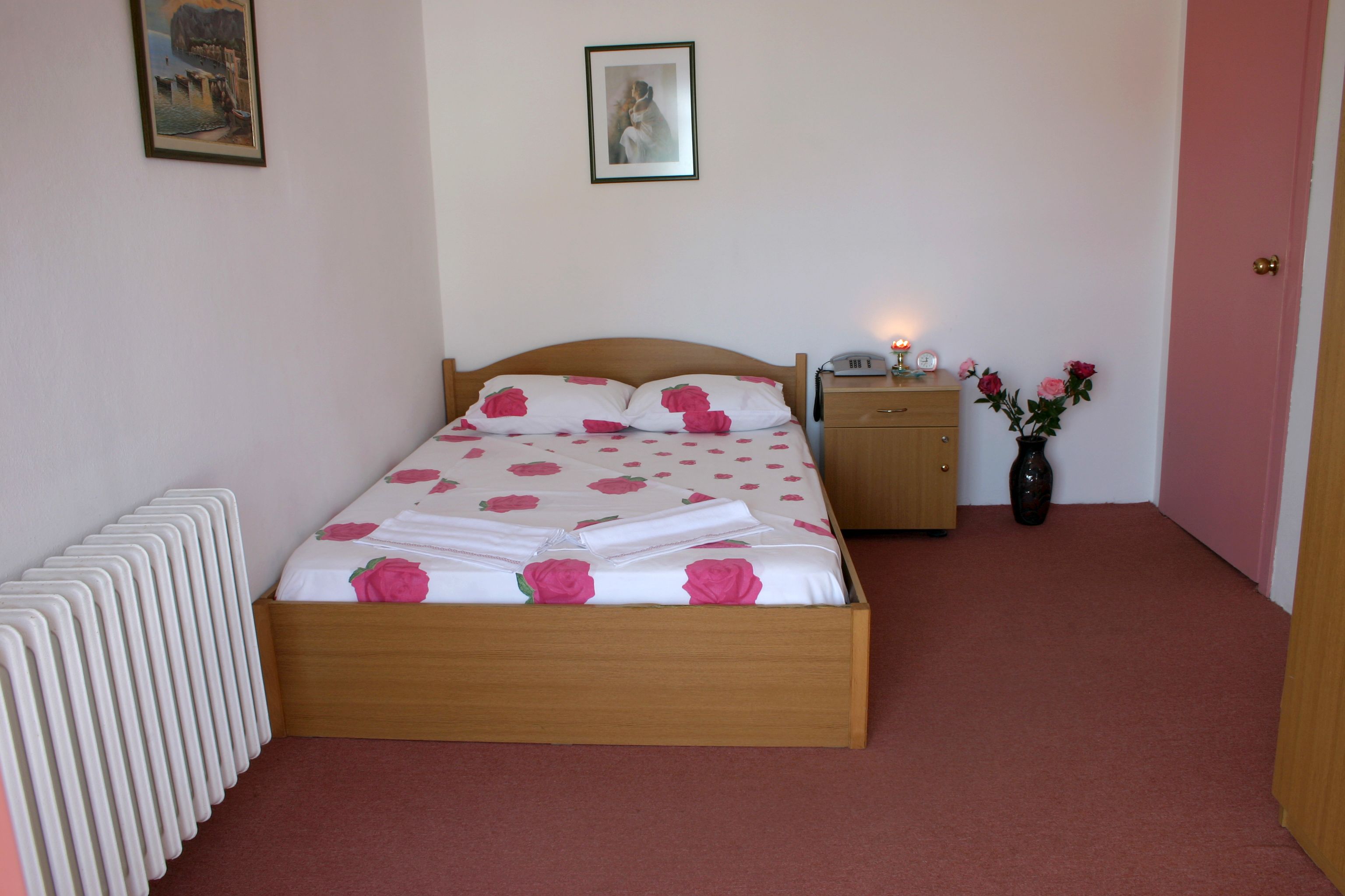 Un dormitorio con una cama de madera, almohadas blancas, una mesita de noche con una lámpara y un jarrón con flores.