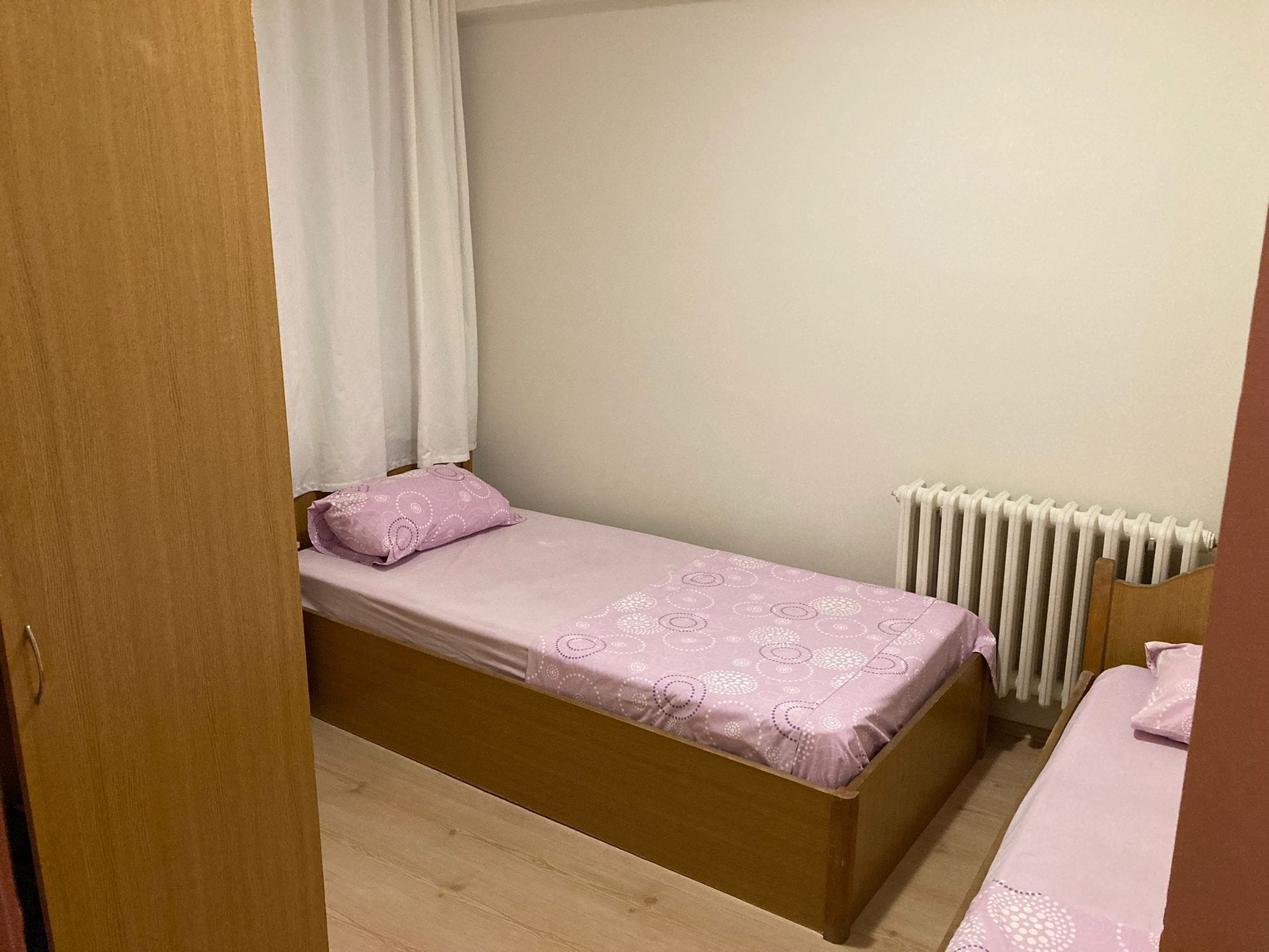 Una habitación pequeña con una cama individual, estructura de madera, radiador blanco y puerta de madera.