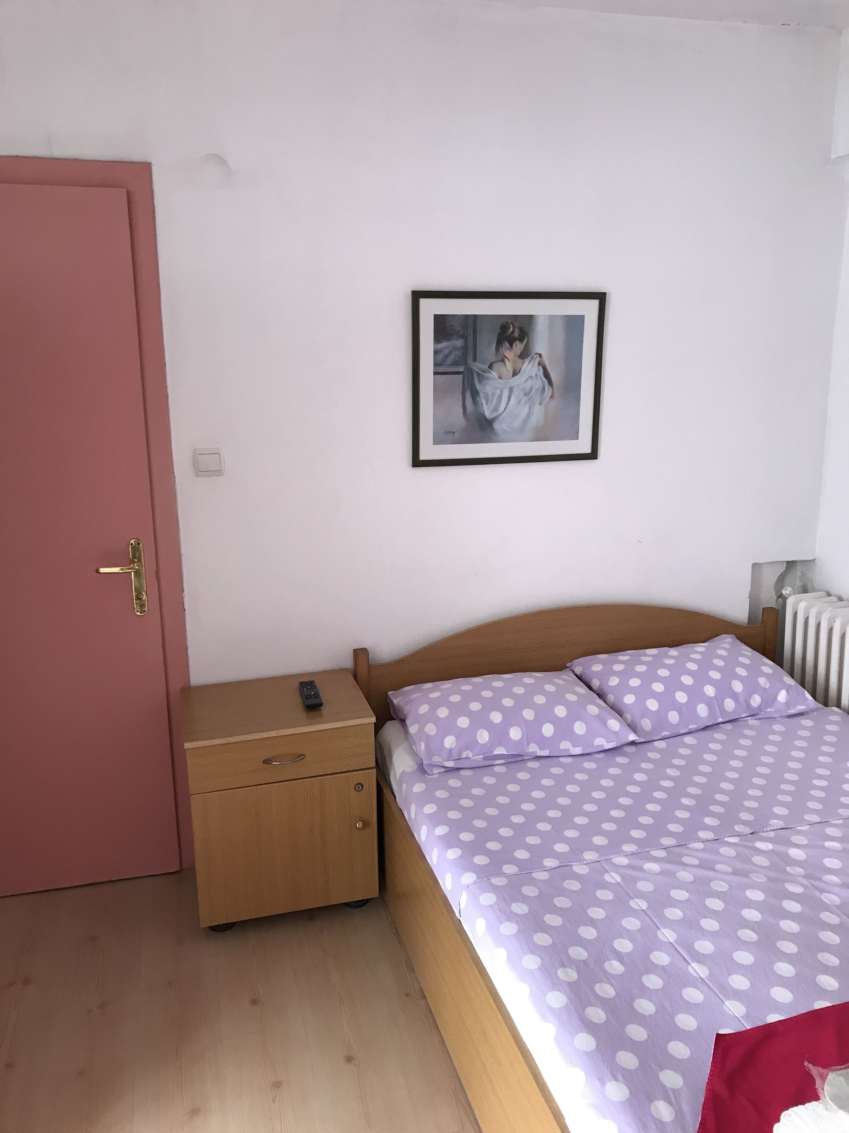 Une chambre avec un lit en bois, une table de chevet, une porte rose, un radiateur blanc et un tableau encadré au mur.