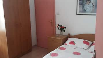A bedroom with a wooden wardrobe, a pink door, a bedside table with a vase of flowers, a bed with a floral bedspread, and a framed picture on the wall.