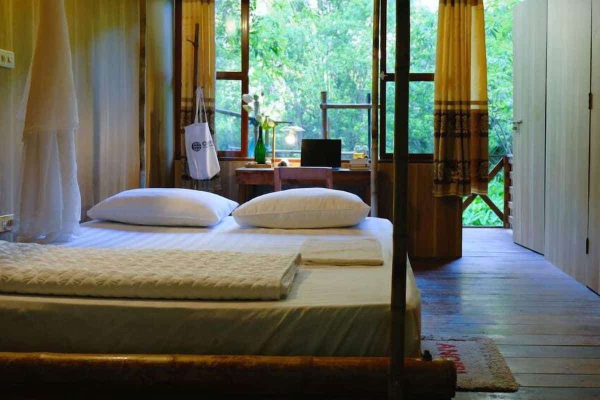 A bedroom with a bed, a desk, and a view of greenery through large windows.